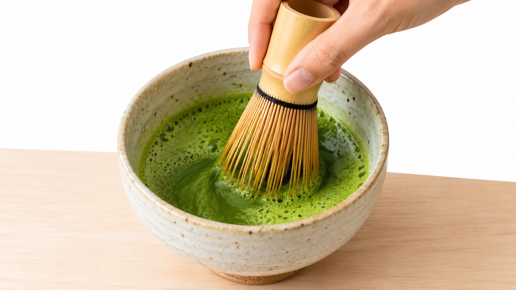 A hand using a bamboo whisk to mix vibrant green matcha tea in a ceramic bowl